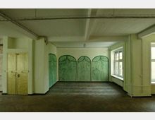 Ein leerer Raum mit hellgrünen Wänden, einer gemalten Wanddekoration aus vier grünen Bögen mit Pflanzenmotiven und Industriestilelementen. Links befindet sich eine goldene Metalltür, rechts zwei Fenster, die Tageslicht hereinlassen.