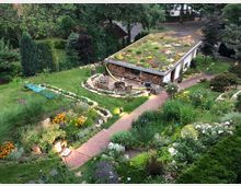 Garten mit einem begrünten Gartenschuppen und Sitzbereich. Die Umgebung ist reich bepflanzt mit Blumen, Büschen und Gehwegen aus rotem Pflasterstein, eingerahmt von Rasenflächen und Bäumen im Hintergrund.