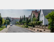 Ruhige Wohnstraße mit Einfamilienhäusern, gepflegten Vorgärten und Gehwegen, umgeben von Bäumen und Sträuchern; im Hintergrund ein mehrstöckiges Gebäude. Ein Fußgänger und parkende Autos sind zu sehen.