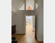 Ein heller Innenraum mit Holzboden und weiß gestrichenen Wänden. Durch eine geöffnete Tür führt der Blick in einen weiteren Raum mit hohen Decken und großen Fenstern, die ein dreieckiges Holzdetail im oberen Bereich einrahmen.