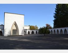 Weiß verputztes Gebäude mit spitzbogigem Eingang und angrenzenden Arkadengängen in einem offenen Hof; im Hintergrund sind Bäume und ein klarer blauer Himmel erkennbar.
