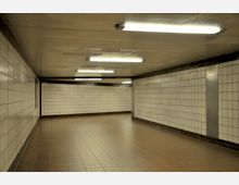 Unterführung mit weißen, gefliesten Wänden und braunem Fliesenboden. Die Decke ist mit mehreren Leuchtstofflampen beleuchtet, und der Raum wirkt schlicht und funktional.