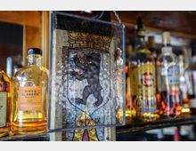 Nahaufnahme einer Barablage mit verschiedenen Flaschen alkoholischer Getränke im Hintergrund. Im Vordergrund ist ein dekoratives Glas mit dem Berliner Wappen und einem schwarzen Bären sowie der Aufschrift „Berlin“ sichtbar.