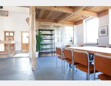 Heller Raum mit Holzdecke und Holzelementen, ausgestattet mit einem langen Holztisch und Stühlen mit orangefarbenen Polstern. Große Fenster lassen Tageslicht hinein, und auf der rechten Seite sind eine Zimmerpflanze und ein schwarzes Regal sichtbar.