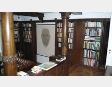 Holzgetäfelte Bibliothek mit dunklen Holzsäulen, deckenhohen Bücherregalen und einem zentral platzierten Kunstwerk, das ein abstrahiertes Gesicht zeigt. Im Vordergrund steht ein Ledersofa neben einem kleinen Tisch mit Büchern und Dekorationen.