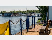 Bootsanleger mit mehreren festgemachten Booten auf einem See, umgeben von grünem Uferbewuchs. Im Vordergrund befinden sich eine Holzbank, ein Geländer mit Ketten sowie eine gelbe Plane.