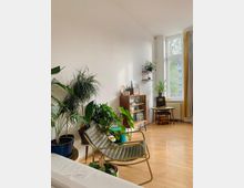 Helles Wohnzimmer mit Holzboden, einer hohen weißen Wand und großen Fenstern, die natürliches Licht hereinlassen. Der Raum ist mit vielen Zimmerpflanzen, einem olivgrünen Stuhl, einem hölzernen Schrank, einem kleinen runden Tisch und warmen Dekorationen gestaltet.