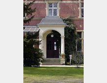 Historische Backsteinfassade eines Gebäudes mit weißem Eingangsportal, das von zwei Säulen getragen wird. Der Eingangsbereich umfasst eine dunkle Holztür mit Glasfenstern, umgeben von Rankpflanzen, und befindet sich in einer Gartenanlage mit gepflegtem Rasen und Bäumen.