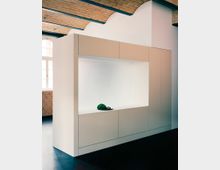 Minimalistischer Innenraum mit einer modernen Einbaustruktur aus weißen Schränken, die eine vertiefte Nische enthalten. Im Hintergrund sind eine Ziegelsteinwand, ein rundbogiges Fenster und eine gewölbte Ziegeldecke erkennbar.