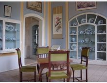 Ein Raum mit historischen Möbeln, darunter ein Holztisch und vier grün gepolsterte Stühle, umgeben von Vitrinen mit blau-weißem Porzellan. Die hellblauen Wände sind mit Gemälden und einem Rollbild dekoriert, und ein offener Durchgang führt zu einem weiteren Raum.