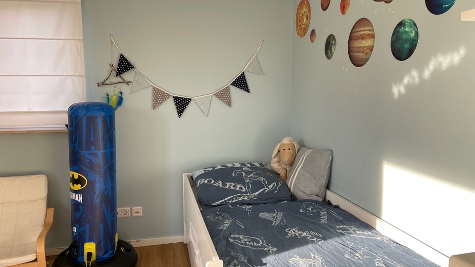 Ein Kinderzimmer mit hellblauen Wänden, dekoriert mit Wandstickern von Planeten und kleinen Sternen. Links steht ein aufblasbarer blauer Boxsack mit Batman-Motiv, daneben ein Bett mit blauer Bettwäsche und einem Kuscheltier, sowie eine weiße Lamellenjalousie am Fenster.