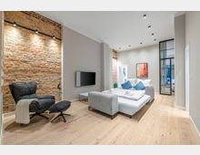 Modernes, helles Schlafzimmer mit freiliegender Backsteinwand, hohen Decken und hellem Holzboden. In der Mitte steht ein graues Polsterbett mit Kissen; daneben sind ein schwarzer Fernseher an der Wand, ein Sessel mit Hocker und eine Glas-Metall-Schiebetür zu einem Nebenraum.