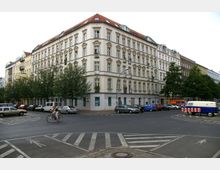 Ecke eines Wohngebäudes im Altbaustil mit hell verputzter Fassade, zahlreichen Fenstern und roten ziegelgedeckten Dächern, umgeben von parkenden Autos und vereinzelten Bäumen. Im Vordergrund eine Straßenkreuzung mit gekennzeichneten Zebrastreifen, ein Fahrradfahrer sowie straßentypische Verkehrsmarkierungen.