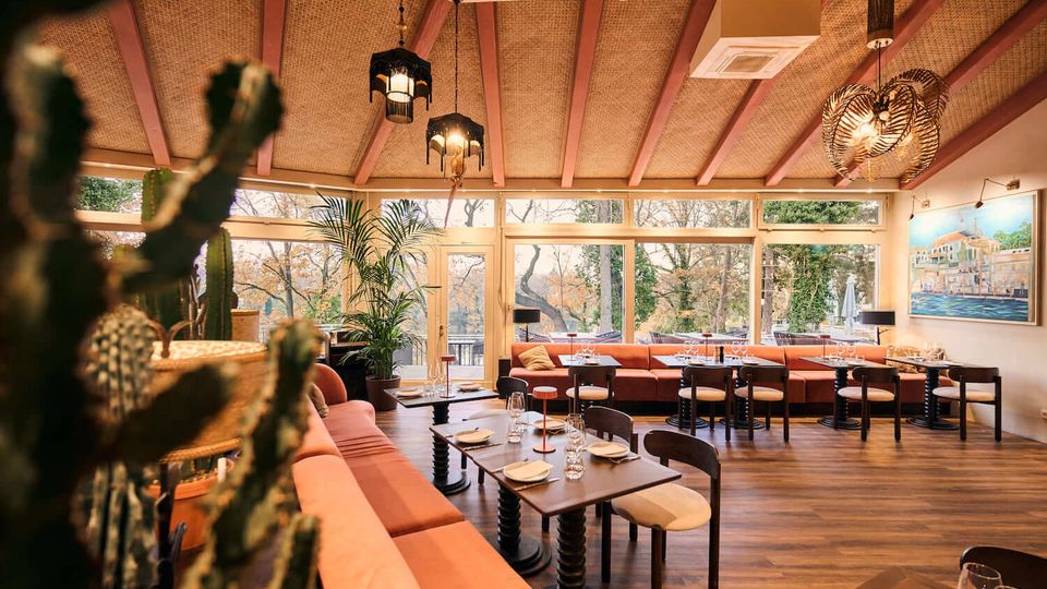 Warm, modern restaurant dining room with long upholstered banquette seating, dark wood tables set with glassware, and pendant lights hanging from a beamed ceiling. Floor-to-ceiling windows overlook trees outside, with potted plants and a large framed wall painting adding decor.