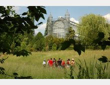 Eine große historische Glaskonstruktion eines Gewächshauses im Hintergrund, umgeben von Bäumen und Sträuchern. Im Vordergrund steht eine Gruppe von Menschen auf einer weitläufigen Wiese, teils von Blattwerk eingerahmt.