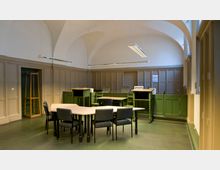 Ein historischer Raum mit hohen Gewölbedecken, Holzvertäfelung und schlichten Leuchten. Im Vordergrund stehen ein Tisch mit Stühlen sowie zwei grüne, erhöhte Rednerpulte; im Hintergrund sind weitere grüne Möbel sowie eine Garderobe zu sehen.