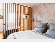 Schlafzimmer mit einer Trennwand aus vertikalen Holzlatten, die zwei Bereiche des Raums abteilt. Im Vordergrund ist ein Bett mit blaugrauer Bettwäsche, im Hintergrund eine helle Wand mit floralen Mustern und ein Fenster, das Licht hereinlässt.
