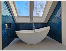 Modernes Badezimmer mit freistehender weißer Badewanne auf einem glatten Boden. Die Rückwand ist mit dunkelblauen Fliesen in Fischgrätenmuster verkleidet, darüber befinden sich große Dachfenster, die einen Blick auf den Himmel und kahle Äste zeigen.