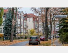 Straßenszene in einem Wohngebiet mit mehrstöckigen, historischen Wohnhäusern im Gründerzeitstil, gekennzeichnet durch rote Dachziegel, Erker und Balkone. Entlang der Pflasterstraße stehen geparkte Autos, während Herbstbäume mit gelbem und braunem Laub den Straßenrand säumen; ein Straßenschild mit "Großbeerenstraße" ist sichtbar.