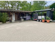 Ein einfacher Holzschuppen mit überdachter Veranda, auf der Gegenstände wie Stühle, Tische und Werkzeuge stehen, daneben ein Anhänger mit Metallaufbau und Pflanzkübeln. Umgeben von Bäumen und Sträuchern auf gepflasterter Fläche.