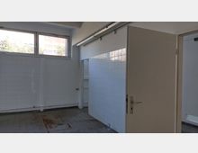 Ein leerer Raum mit weißen Fliesen an den Wänden und grauem Fliesenboden. Oben an der Wand sind Fenster mit Blick auf Bäume und ein Gebäude sichtbar, während eine offene Tür in einen weiteren Raum führt.