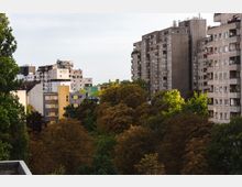Städtische Wohnsiedlung mit mehrstöckigen Wohnhäusern aus Beton, teilweise mit Graffiti an den Fassaden, umgeben von dichtem Baumbestand in Herbstfarben. Im Hintergrund sind weitere Gebäude und ein Turm sichtbar.