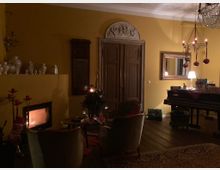 Wohnzimmer mit gelb gestrichenen Wänden, einem Kamin mit brennendem Feuer, einem kleinen geschmückten Weihnachtsbaum und einer antiken Holztür mit dekorativem Relief oben. Im Raum stehen Sessel, ein Tisch, ein Flügel, ein großer Wandspiegel und ein Kronleuchter mit roten Weihnachtskugeln.