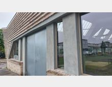 Gebäude mit moderner Stein- und Putzfassade, großen Fenstern und einer Metalltür; im Hintergrund sind Glasdach-Reflexionen, parkende Autos und Grünflächen erkennbar.