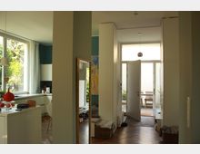 Heller Innenraum einer modernen Wohnung mit offener Küche, Parkettboden und einem großen Fenster, das Tageslicht hereinlässt. Ein offener Flur führt zu einer weißen Haustür, die in einen kleinen Außenbereich mit Holzmöbeln und Pflanzen führt.