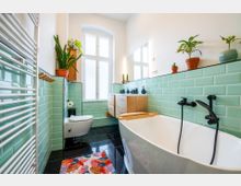 Helles Badezimmer mit mintgrünen Fliesen, freistehender weißer Badewanne mit schwarzen Armaturen, einem Holzwaschbeckenunterschrank und großen Fenstern. Verschiedene Topfpflanzen dekorieren den Raum, der einen modernen und natürlichen Stil hat.