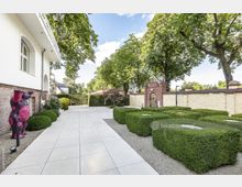 Gepflegter Außenbereich mit gepflastertem Weg, geschnittenen Buchsbaumhecken und kleinen Bäumen, umgeben von einer gelblichen Backsteinmauer. Links ist ein weißes Gebäude mit Gerüstbögen und einer modernen Skulptur sichtbar, im Hintergrund stehen große, schattenspendende Bäume.