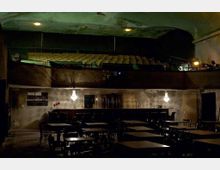 Ein düsterer Innenraum eines ehemaligen Theaters oder Kinos mit einer kleinen Bar im unteren Bereich und Sitzplätzen auf zwei Ebenen, darunter Tische mit Stühlen im Vordergrund und eine Reihe von Sitzreihen auf einer Empore darüber. Die Wände wirken abgenutzt, und ein zentraler Kronleuchter spendet schwaches Licht.