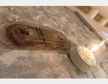 Nahaufnahme einer rustikalen Wand mit sichtbarer Patina und Metallbefestigung, daneben eine montierte elektrische Glühbirne in einem transparenten Gehäuse. Im Hintergrund sind unscharf helle Wand- oder Deckenbereiche zu erkennen.