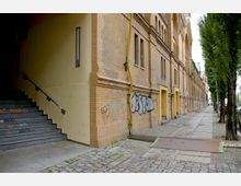 Gelbes Backsteingebäude mit graffitibekritzelten Wänden, breiten Treppen links, sowie einem Gehweg aus Pflastersteinen und Gehplatten rechts, gesäumt von hohen Bäumen.