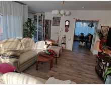 Wohnzimmer mit beigen Ledersofas, Holztischen und einer Mischung aus gemütlicher Dekoration wie Pflanzen, Decken und Kissen. Im Hintergrund ein angrenzender Essbereich mit Holztisch und Stühlen, getrennt durch eine offene Tür mit weißen Vorhängen.