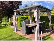 Überdachte Gartensitzecke mit Metallpavillon und seitlichen Vorhängen auf einer Holzterrasse, darunter ein Esstisch mit mehreren geflochtenen Stühlen. Umgeben von Rasen, hohen Hecken, Blumenbeeten und einem kleinen Gartenhaus im Hintergrund.