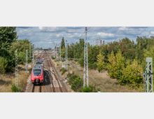 Eine Regionalbahn auf einem mehrgleisigen Bahngleis fährt durch eine grüne, leicht bewaldete Umgebung. Im Hintergrund sind Hochspannungsleitungen, Oberleitungsmasten und ein Wohnhochhaus vor einem bewölkten Himmel zu erkennen.