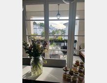 Ein Blick durch große Glastüren auf einen überdachten Balkon mit Sitzmöbeln, Pflanzen und dekorativer Metallgeländer. Im Vordergrund stehen ein Vase mit Blumen sowie mehrere Gläser mit Lebensmittel auf einem Tisch.