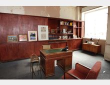 Ein schlicht eingerichtetes Büro im Retro-Stil mit Holzmöbeln, darunter ein Schreibtisch mit grünem Lederbezug, ein roter Sessel und eine große Wandregaleinheit mit Büchern, Dekorationsobjekten und Bildern. Ein großes Fenster rechts lässt Tageslicht in den Raum.