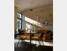 Ein gemütliches Café mit Holzmöbeln und einer orangefarbenen Couch im Hintergrund. An der beigefarbenen Wand hängen Bilderrahmen und Pflanzen, und durch ein großes Fenster fällt Sonnenlicht in den Raum.