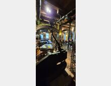 Rustikaler Innenraum eines Restaurants mit Backsteinbögen, sichtbaren Deckenbalken und industriellen Rohren unter der Decke. Im Vordergrund steht eine große schwarze Presse oder Maschine, dahinter Tische und Stühle sowie schmiedeeiserne Kerzenhalter mit roten Kerzen auf Fliesenboden.