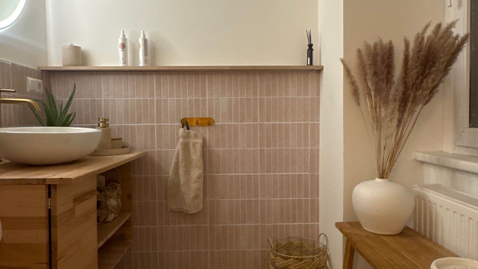 Helles Badezimmer mit halbhoher, beige gefliester Wand und grauem Steinboden. Links steht ein Holzwaschtisch mit runder Aufsatzschale und goldfarbenem Wasserhahn, rechts eine Holzbank neben einem Fenster mit weißer Vase und Trockenpflanzen.