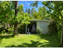 Garten mit grünem Rasen, umgeben von Bäumen und Sträuchern. Im Hintergrund steht ein kleines, helles Gartenhaus mit zwei Türen und einem überdachten Bereich, in dem Gartengeräte und Möbel abgestellt sind.