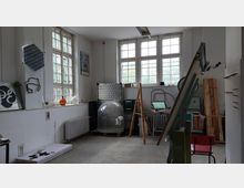 Ein Werkraum mit weißen Wänden, großen Fenstern mit Holzrahmen und verstreuten Gegenständen wie einer Glaskuppel, Geräten, Werkzeugen und Materialien. Im Vordergrund stehen ein Holzgestell und eine rote Stuhl.