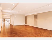 Großer leerer Innenraum mit Holzfußboden, beigen Wänden und deckenintegrierten Einbauleuchten. Im Hintergrund sind Fenster und Glastüren zu sehen, die Tageslicht einlassen.