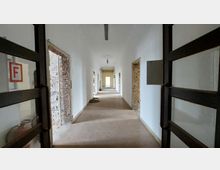 Langer, schmaler Flur in einem renovierungsbedürftigen Gebäude, mit weißen Wänden und teils freigelegtem Mauerwerk an den Türöffnungen. Am Ende des Flurs ist eine geöffnete Tür mit Blick in einen weiteren Raum sichtbar.