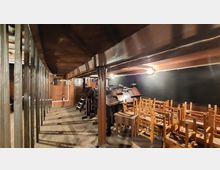 Ein schmaler Raum unter einem hölzernen Deckengewölbe mit Holzstützbalken, bestückt mit Pulten, Stühlen und Tischen. Der Raum wird durch mehrere kleine Lampen beleuchtet, und die Wände sind dunkel gestaltet.