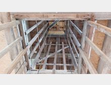 Blick von unten in eine hölzerne Konstruktion mit Gitterverstrebungen, schräg nach oben verlaufenden Balken und sichtbaren Seilen; umgeben von unbehandeltem Holz und OSB-Platten.