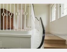 Treppenhaus in einem hellen Innenraum mit Holzstufen und einem weißen Geländer mit schwarzem Handlauf. Im Hintergrund steht ein antiker Holzschrank vor einer offenen Tür sowie ein farbiger Teppich auf dem Boden.