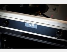 Nahaufnahme eines modernen Einbauherds mit schwarzer Glasfront und Edelstahlblende, mit zentralem Digitaldisplay und Drehknöpfen links und rechts. Oben ist ein Glaskeramik-Kochfeld mit leuchtenden Bedienanzeigen und einem Topf zu sehen.